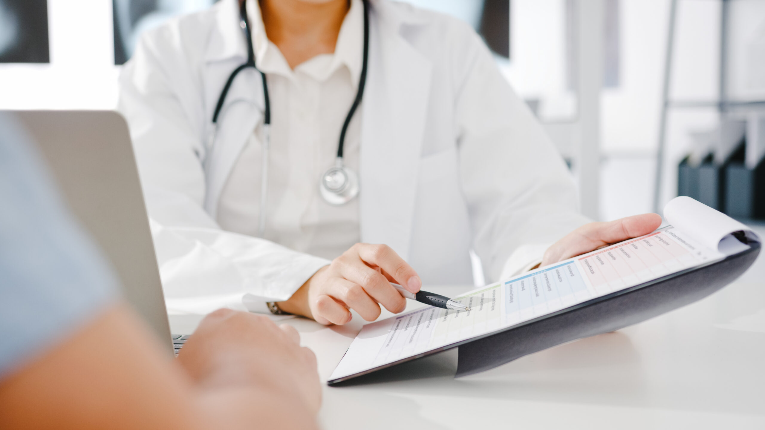 Young Asia female doctor in white medical uniform using clipboard is delivering great news talk discuss results or symptoms with male patient sitting at desk in health clinic or hospital office.